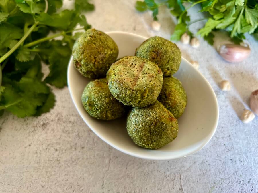 Small dish with falafel balls in it and fresh cilantro and parsley in the background