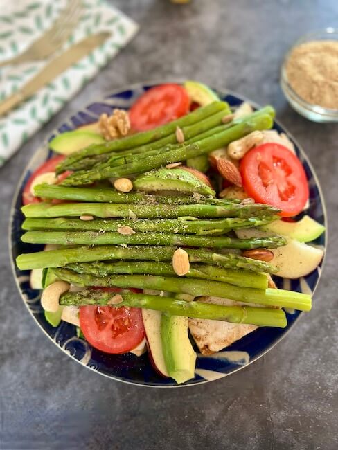 Favourite way to eat asparagus: bowl of salad, green leaves, cucumber, red pepper, sliced tomato, tofu, avocado, apple and asparagus spears on top sprinkled with mixed nuts
