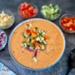 Bowl of mango gazpacho with toppings of red pepper, tomato, onion, cucumber and apple
