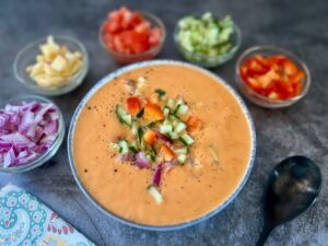 Bowl of mango gazpacho with toppings of red pepper, tomato, onion, cucumber and apple