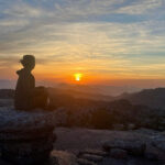 Stay grounded while manifesting - person sitting on a rock staring out to the distant sunsest over the mountains
